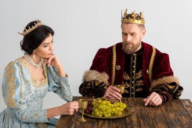 thoughtful queen and king with crowns sitting at table isolated on grey