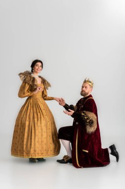 king with crown bending on knee and holding hand of smiling queen on grey background 
