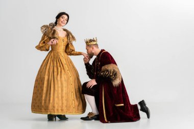 king with crown bending on knee and kissing hand of smiling queen on grey background 