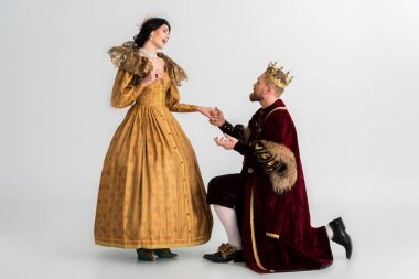 king with crown bending on knee and holding hand of shocked queen on grey background 
