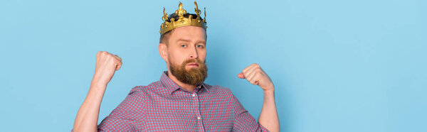 panoramic shot of serious man with crown showing strong gesture on blue background 