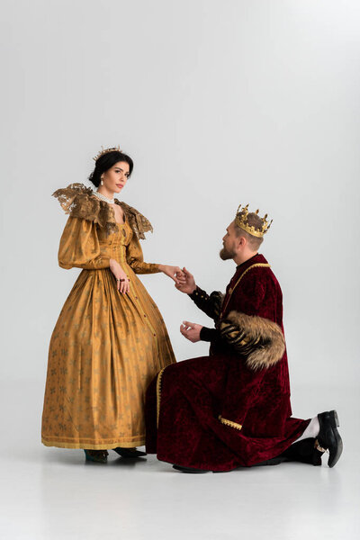 king with crown bending on knee and holding hand of queen on grey background 