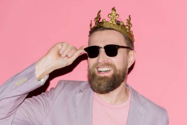 smiling man with crown touching sunglasses on pink background 