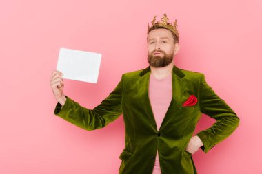 handsome man in velour jacket and crown holding digital tablet on pink background 