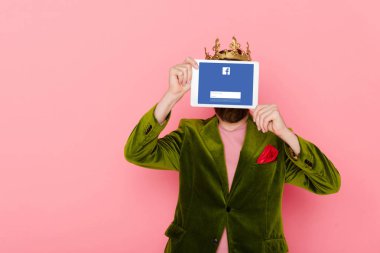 KYIV, UKRAINE - DECEMBER 3, 2019: man with crown holding digital tablet with facebook app isolated on pink