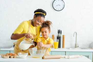 Neşeli Afrikalı Amerikalı anne mutfaktaki tatlı kızın yanına süt döktü. 