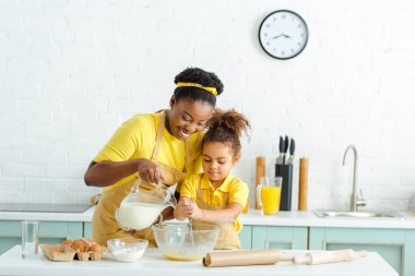 cheerful african american mother pouring milk in bowl near cute kid  clipart