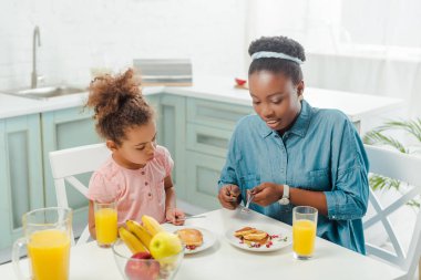 Afro amerikan anne ve kızı ellerinde çatal bıçak takımı ile lezzetli gözlemelerin yanında tabakta 