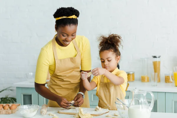 陽気なアフリカ系アメリカ人の母親とかわいい娘でエプロン彫刻生餃子でキッチン — ストック写真