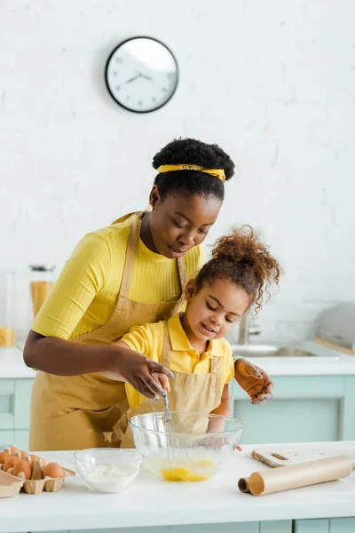 かわいいアフリカ系アメリカ人の子供と幸せな母親は台所で料理をしながらボウルの近くに泡立て器を保持 — ストック写真