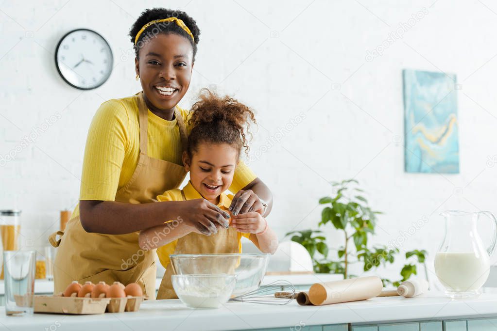 cute african american kid and cheerful mother adding egg in bowl while cooking in kitchen 