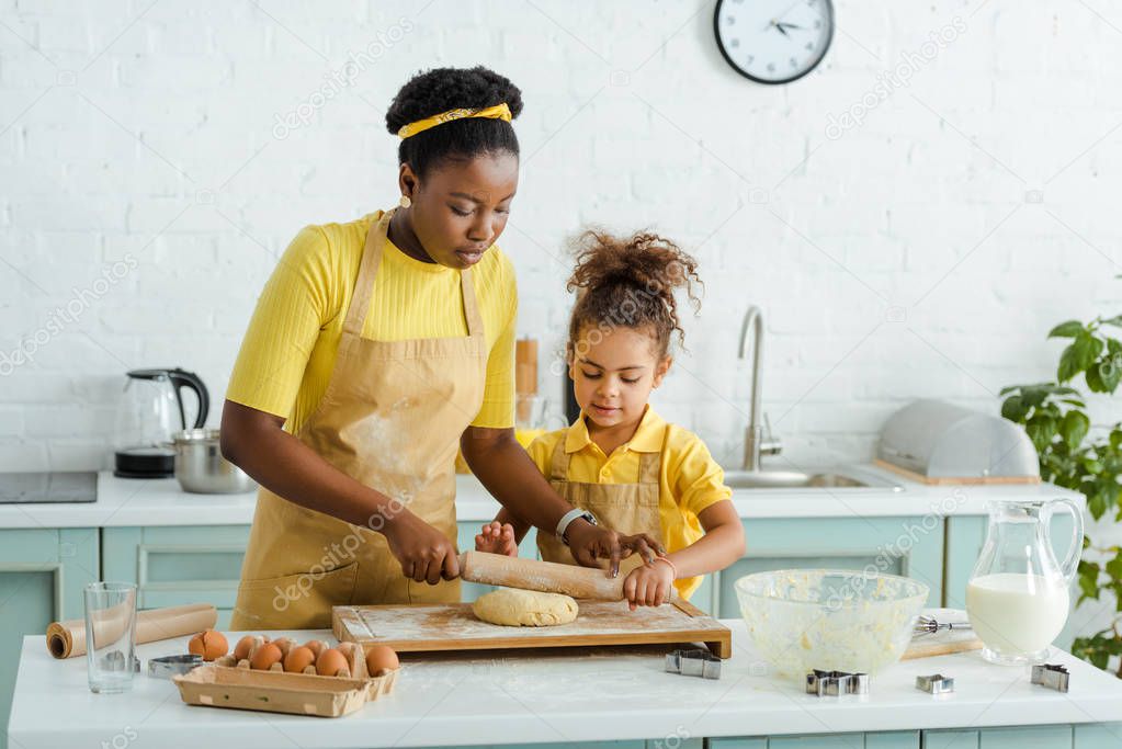cute african american daughter looking at raw dough near mother holding rolling pin 