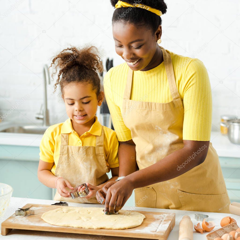 adorable african american kid and happy mother holding cookie cutters near raw dough 
