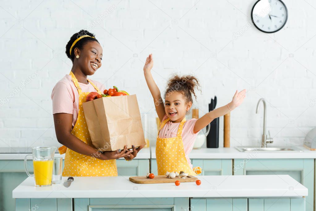 happy african american mother holding paper bag with groceries near cheerful daughter with hands above head 