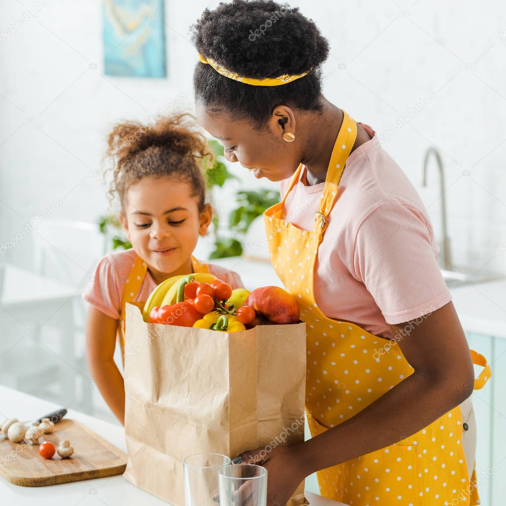 happy african american mother and daughter looking at paper bag with groceries 