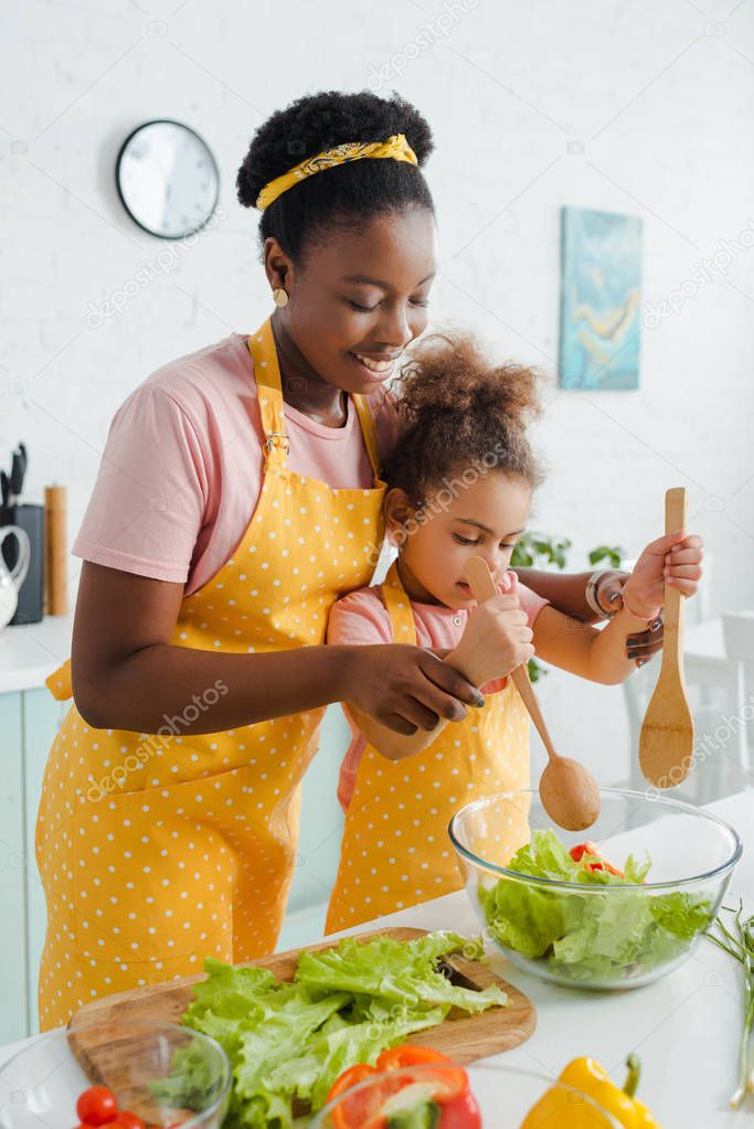 cheerful african american mother and cute child holding wooden spoon and spatula while preparing salad 