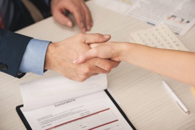 cropped view of agent and client shaking hands near clipboard with insurance lettering  clipart