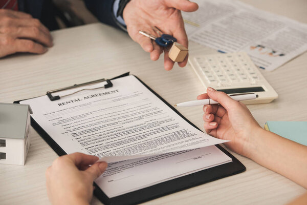 cropped view of woman holding pen near clipboard with rental agreement lettering and agent with keys 