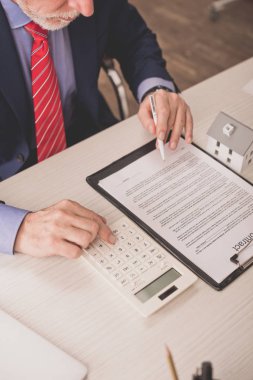 cropped view of bearded broker using calculator and holding pen near clipboard  clipart