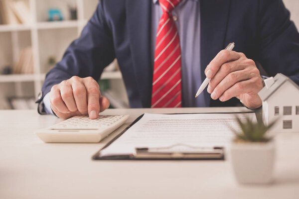 selective focus of broker using calculator and holding pen near clipboard and plant 