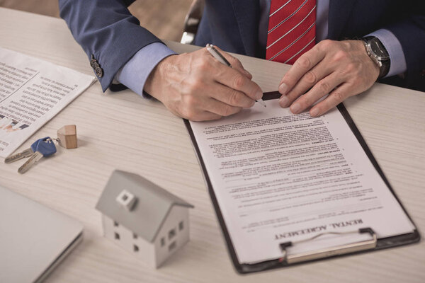 cropped view of agent holding pen near clipboard with rental agreement, carton house model and keys on desk 