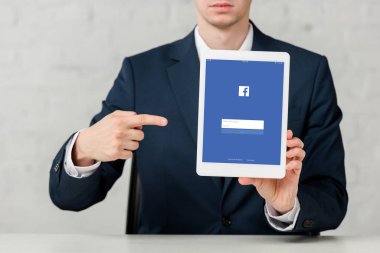 cropped view of realtor in suit pointing with finger at digital tablet with facebook app on white 