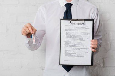 cropped view of realtor in suit holding key and clipboard with lease agreement on white 