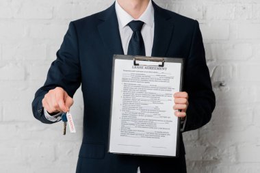 cropped view of realtor in suit holding key with leasing lettering and clipboard with lease agreement on white  clipart