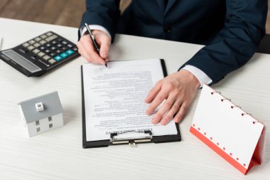 cropped view of realtor signing lease agreement near blank calendar, calculator and house model