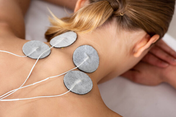 Back view of woman with electro stimulation electrodes on neck on massage couch isolated on grey