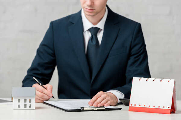 cropped view of realtor writing near blank calendar and house model on white, leasing concept 