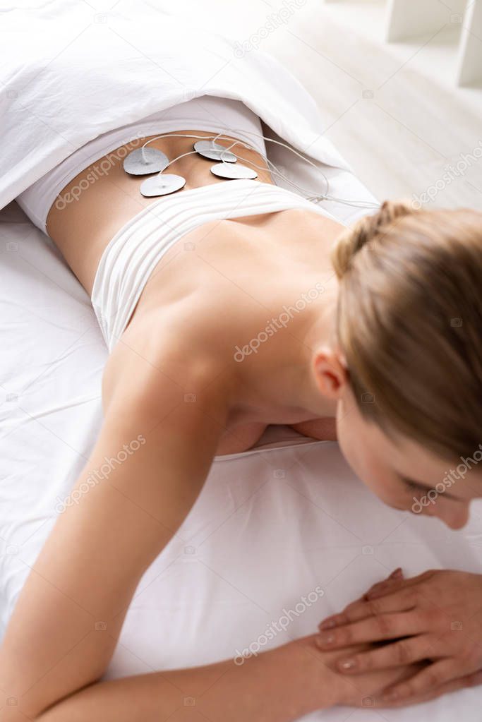 Overhead view of young woman during electrode treatment of lower back on massage couch in clinic