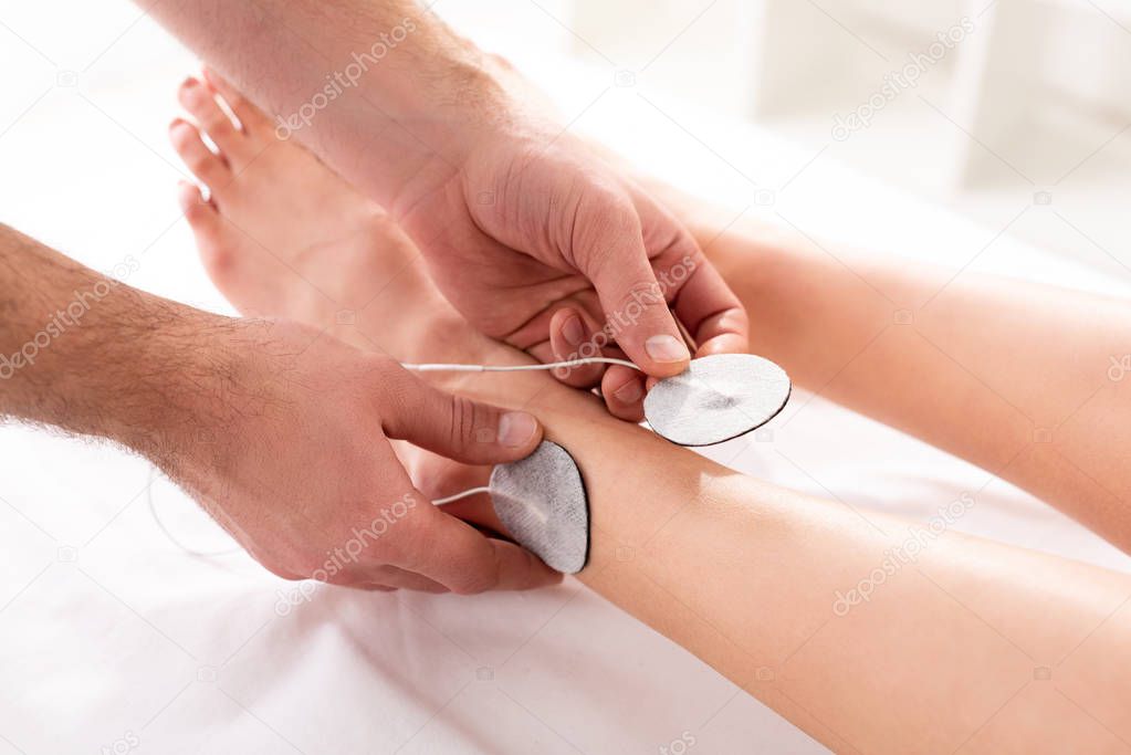 Cropped view of therapist setting electrodes on leg of patient during electrotherapy in clinic