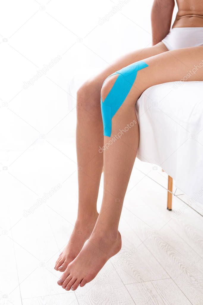 Cropped view of woman with kinesiology tapes on knee sitting on massage couch on white background