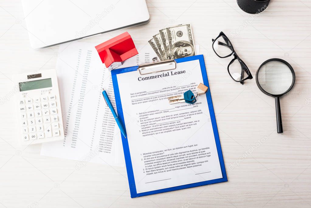 top view of document with commercial lease lettering on clipboard near keys, house model, pen, calculator, magnifier, money and glasses