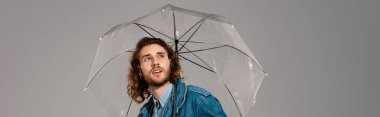 Gri renkli bir şemsiye tutan, kot ceketli yakışıklı ve şok olmuş bir adamın panoramik çekimi.