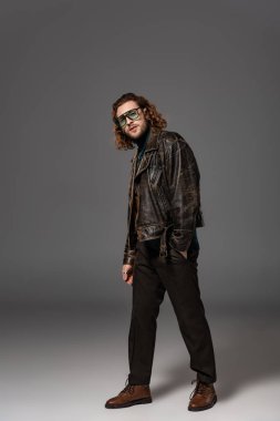 Deri ceketli ve pantolonlu yakışıklı adam gri arka planda kameraya bakıyor. 