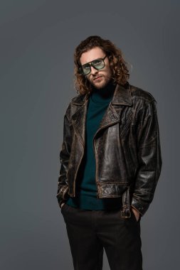 Deri ceketli yakışıklı, elleri ceplerinde gri renkli kameraya bakıyor.