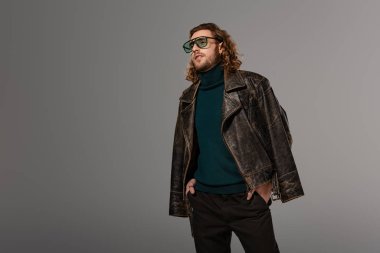 Deri ceketli yakışıklı adam, elleri ceplerinde. Gri renkten başka yere bakıyor.