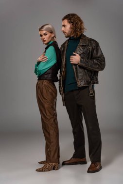 Deri ceketli bir adam gri arka planda çapraz kollu bir kadına bakıyor. 