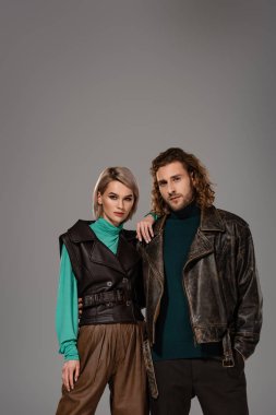 Yelekli kadın ve deri ceketli adam gri kameraya bakıyor.