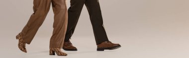 Pantalon ve ayakkabı giyen kadın ve erkeğin gri arka planda yürüdüğü panoramik bir fotoğraf. 