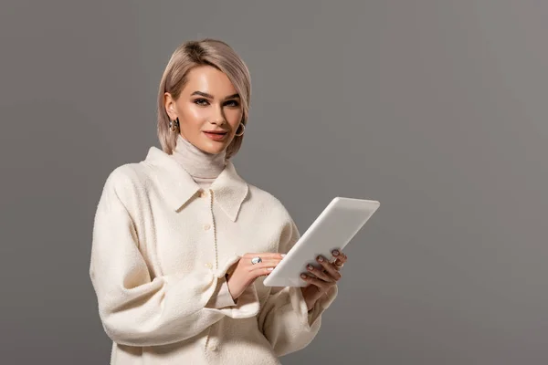 グレーで隔絶されたデジタルタブレットを持つ白いコートの魅力的で笑顔の女性 — ストック写真