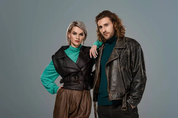 woman in vest and man in leather jacket looking at camera isolated on grey