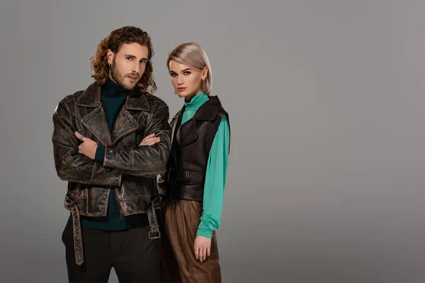 woman in vest and man in leather jacket with crossed arms looking at camera isolated on grey