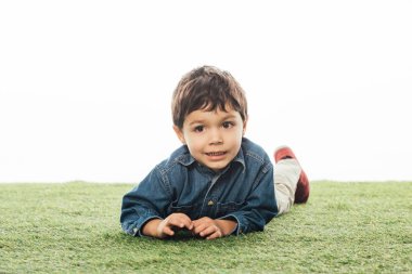 cute and smiling boy looking away and lying on grass isolated on white 