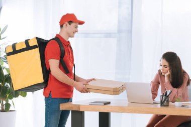 Sırt çantalı gülümseyen kurye masada iş kadınının yanında pizza kutuları tutuyor.