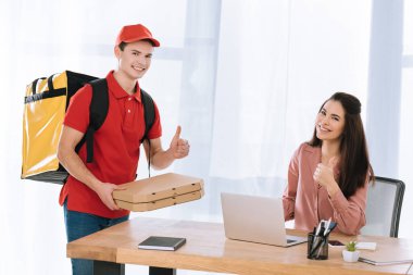 Pizza kutularıyla teslimatçı masadaki iş kadınının yanında baş parmak işareti gösteriyor.