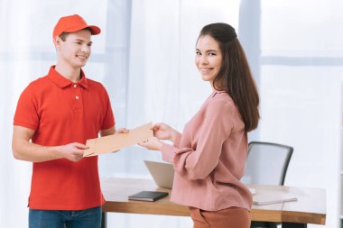 Courier giving envelope to smiling businesswoman in office clipart
