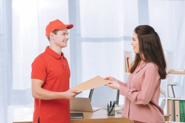 Side view of delivery man giving envelope to smiling businesswoman in office  clipart
